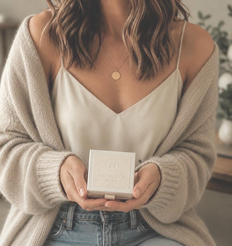 May include: A square, off-white jewelry box with a small pull tab, featuring the embossed letter N inside a circle and the text NICCOLETTE EST. 2018. The box is held in place, accompanied by a gold-toned circular pendant necklace and a beige cardigan.