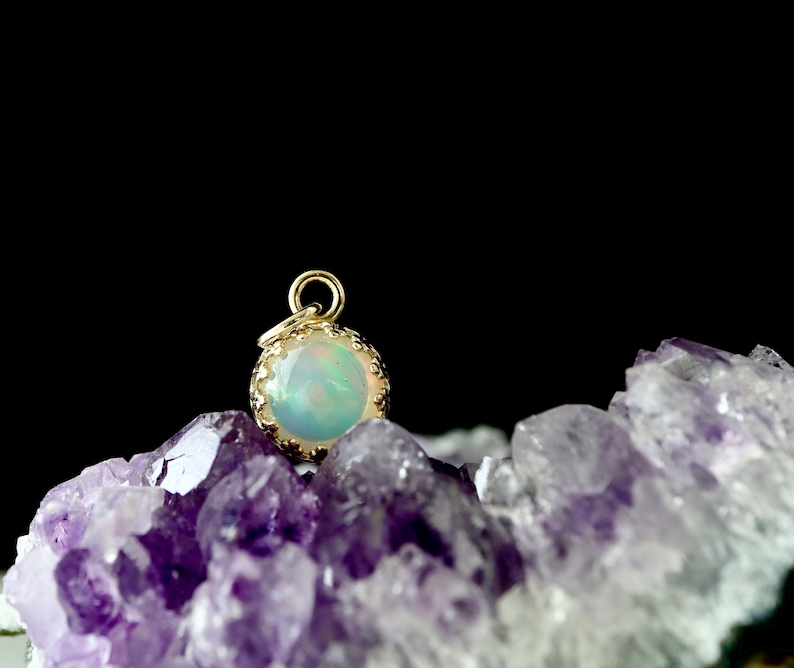 May include: A gold pendant with a round opal gemstone set in a gold bezel. The pendant is sitting on a bed of purple amethyst crystals.