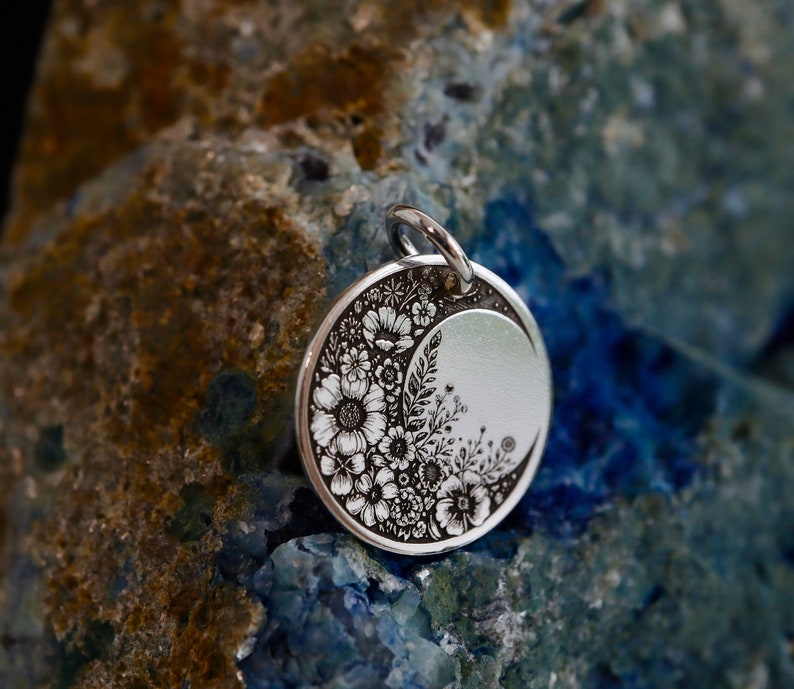 May include: A circular silver pendant featuring a detailed black engraved floral design that curves along one side, leaving a smooth, plain crescent-shaped area on the right. The pendant includes a small silver jump ring at the top for attachment.
