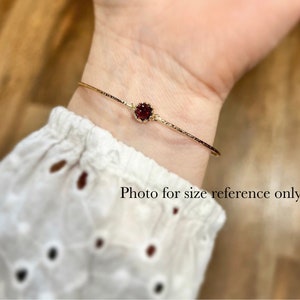 Pode incluir: Um bracelete de ouro com uma &uacute;nica pedra preciosa vermelha engastada no centro. O bracelete &eacute; mostrado no pulso de uma pessoa. "Foto apenas para refer&ecirc;ncia de tamanho" est&aacute; escrito abaixo do bracelete.