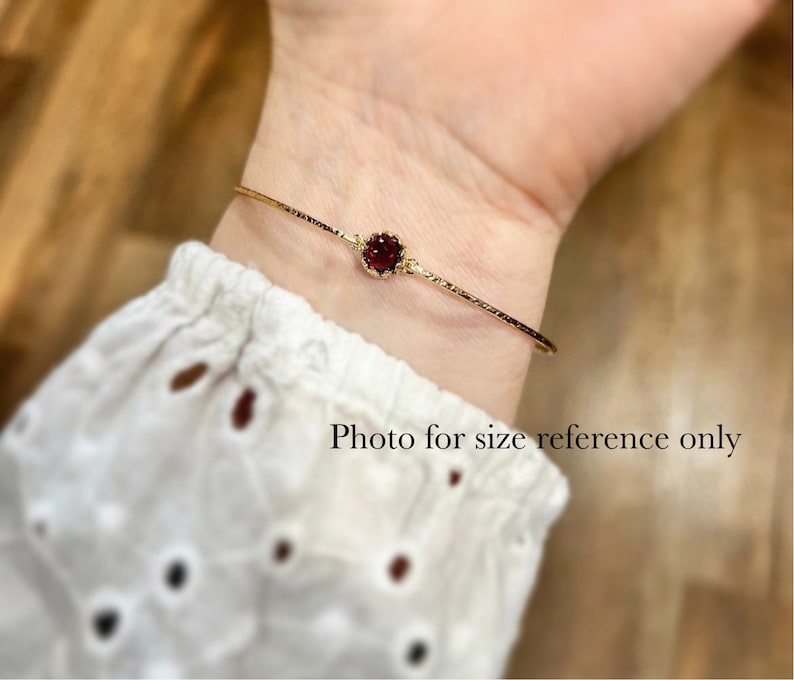 Pode incluir: Um bracelete de ouro com uma &uacute;nica pedra preciosa vermelha engastada no centro. O bracelete &eacute; mostrado no pulso de uma pessoa. "Foto apenas para refer&ecirc;ncia de tamanho" est&aacute; escrito abaixo do bracelete.