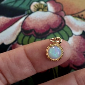 Puede incluir: Un peque&ntilde;o colgante redondo con una piedra de &oacute;palo iridiscente central engastada en un marco de metal dorado con un borde festoneado decorativo y una peque&ntilde;a anilla circular en la parte superior para colgarlo.