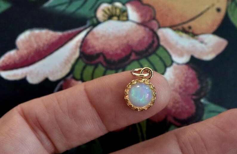Puede incluir: Un peque&ntilde;o colgante redondo con una piedra de &oacute;palo iridiscente central engastada en un marco de metal dorado con un borde festoneado decorativo y una peque&ntilde;a anilla circular en la parte superior para colgarlo.