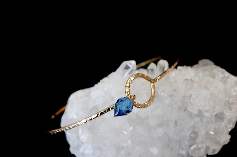 May include: A gold-toned metal bangle bracelet featuring a hammered texture, a circular open link, and a single teardrop-shaped blue gemstone pendant. The bracelet is resting on a cluster of translucent white quartz crystals against a black background.