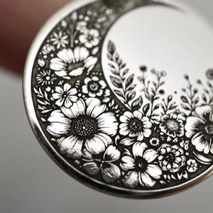 May include: A circular silver pendant featuring a detailed engraved crescent moon shape filled with various floral patterns including daisies and leafy stems. The metal surface has a polished finish with dark recessed lines highlighting the intricate botanical design.