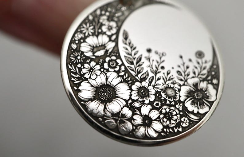 May include: A circular silver pendant featuring a detailed engraved crescent moon shape filled with various floral patterns including daisies and leafy stems. The metal surface has a polished finish with dark recessed lines highlighting the intricate botanical design.