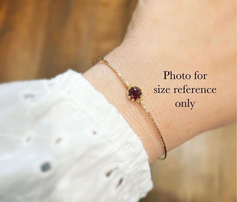 Puede incluir: Un brazalete dorado con una superficie texturizada y una gema roja redonda. El brazalete se muestra en una mu&ntilde;eca, con el texto "Photo for size reference only" visible.