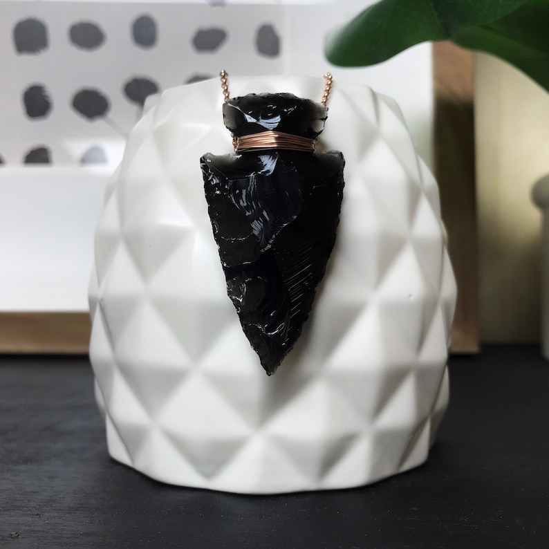 Op de afbeelding: Een ketting met een zwarte obsidianen pijlpunt hanger, een ros&eacute;gouden ketting en een draadwikkeling. De hanger wordt gepresenteerd op een witte keramische standaard.