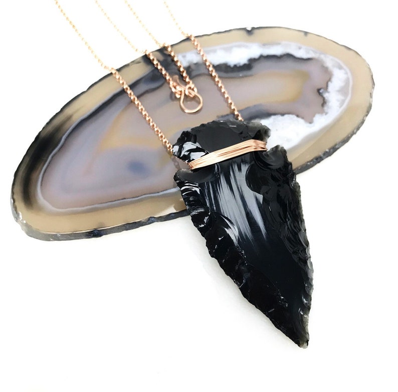 Op de afbeelding: Een zwarte obsidianen pijlpunt hanger met een ros&eacute;gouden draadwikkeling, hangend aan een ros&eacute;gouden ketting op een lichtbruine agaat plak.