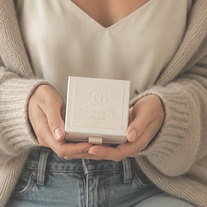 Puede incluir: Un joyero cuadrado de color blanco roto con una peque&ntilde;a leng&uuml;eta en la parte inferior. La tapa presenta un logotipo circular en relieve con la letra N en el centro, seguido del texto PICCOLETTI y el texto m&aacute;s peque&ntilde;o EST. 2018.