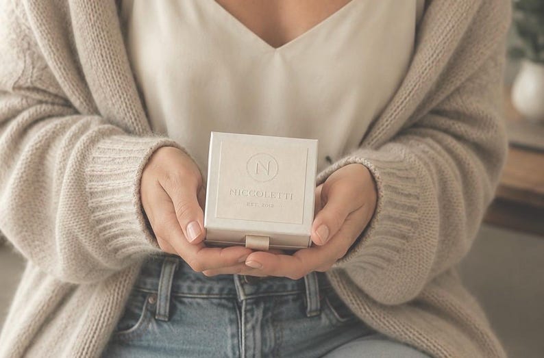 Puede incluir: Un joyero cuadrado de color blanco roto con una peque&ntilde;a leng&uuml;eta en la parte inferior. La tapa presenta un logotipo circular en relieve con la letra N en el centro, seguido del texto PICCOLETTI y el texto m&aacute;s peque&ntilde;o EST. 2018.