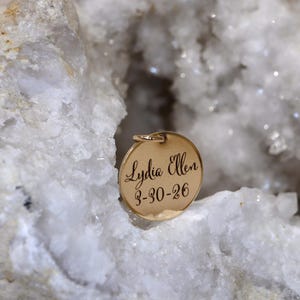 May include: A circular gold-toned metal pendant featuring the engraved name Lydia Ellen and the date 3-30-26 in a script font. The pendant is positioned within the hollow center of a large, rough, white and translucent crystalline rock formation.