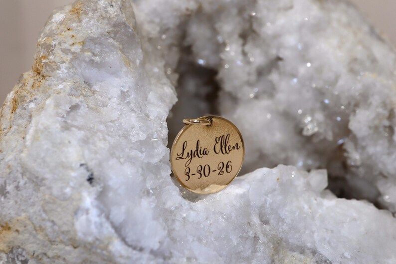 May include: A circular gold-toned metal pendant featuring the engraved name Lydia Ellen and the date 3-30-26 in a script font. The pendant is positioned within the hollow center of a large, rough, white and translucent crystalline rock formation.
