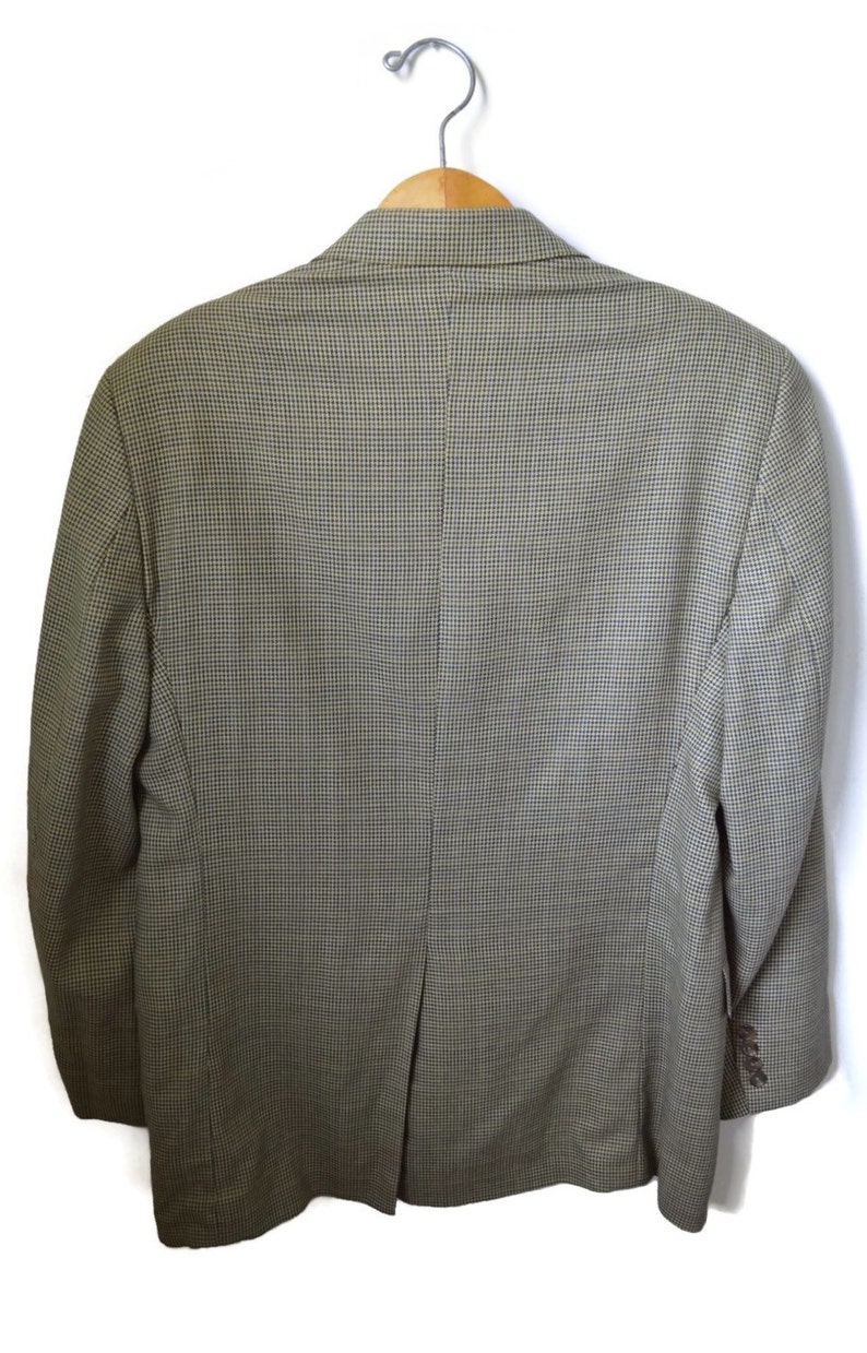 May include: A blazer featuring a fine-scale houndstooth pattern in shades of olive green and brown. The garment is displayed from the back, showing a center vent, long sleeves with four buttons on each cuff, and a structured collar on a hanger.