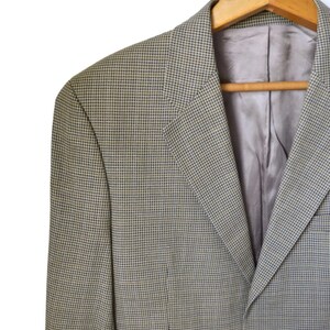 May include: A blazer featuring a fine houndstooth pattern in shades of olive green and dark blue. The garment has notched lapels, a smooth light gray interior lining, and two dark brown buttons on the front, all displayed on a wooden hanger.