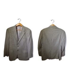 May include: A gray and black micro-check blazer featuring a notched lapel, two front buttons, two flap pockets, and long sleeves. The garment is displayed on a wooden hanger, showing both the front view with a gray lining and the back view.