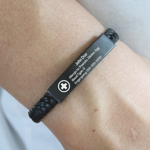 May include: Black leather bracelet with a black metal plate engraved with medical information. The plate includes the name "John Doe", blood type "O", allergies to peanuts, gluten, and egg, and an emergency phone number.
