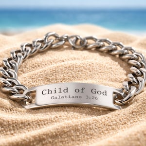 May include: A silver-colored chain bracelet with a rectangular plate engraved with "Child of God" and "Galatians 3:26". The bracelet is resting on sand with a blurred ocean background.