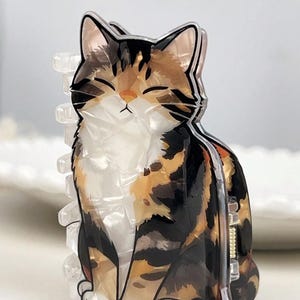 May include: A cat-shaped hair claw clip. The clip features a calico cat design with black, brown, and white markings. The cat is sitting with its eyes closed. The clip is made of clear and opaque plastic.