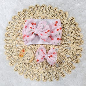 Peut inclure: Deux nœuds pour cheveux roses avec un imprimé cerise. Les nœuds sont sur un fond blanc avec le texte "Little Dahlia Boutique".