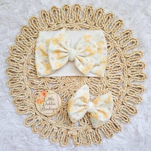 May include: A set of baby headbands and bows. The headbands and bows are cream-colored with a yellow bow pattern and small floral accents. The items are arranged on a woven, circular placemat. A wooden tag with the text "Little Dahlia Boutique" is also present.
