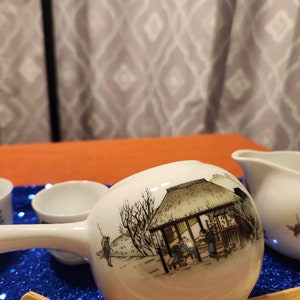May include: White ceramic tea set featuring a long-handled water dipper with a detailed illustration of a traditional Japanese scene. The set includes small cups and a creamer, all with matching designs. The dipper is approximately 8 inches long.