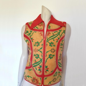 May include: A vintage vest with a vibrant floral pattern. The vest features a tan base with red trim and a red collar. Green and red floral designs adorn the front. The vest has a full-length zipper.