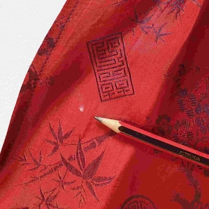 May include: Close-up of red fabric with a dark pattern of leaves and geometric shapes. A pencil with black and red stripes rests on the fabric, pointing towards the center. The fabric has a slight sheen.
