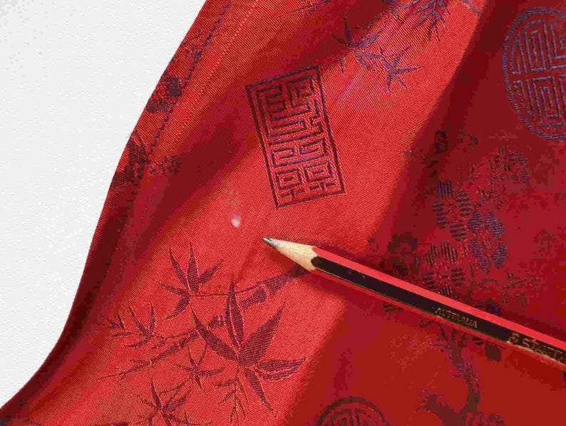 May include: Close-up of red fabric with a dark pattern of leaves and geometric shapes. A pencil with black and red stripes rests on the fabric, pointing towards the center. The fabric has a slight sheen.