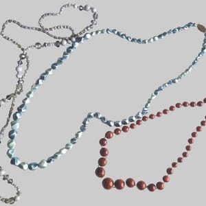 Könnte beinhalten: Drei Halsketten sind abgebildet: eine lange Kette aus hellblauen facettierten Perlen, eine lange Kette aus gemischten weißen, grauen und klaren Kristallperlen sowie eine kürzere Kette aus runden, polierten, rotbraunen Perlen mit Metallic-Finish.