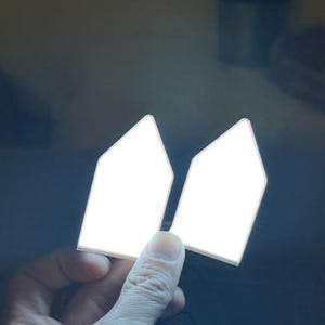May include: Two white, geometric-shaped objects with a bright, glowing appearance. The objects are held by a hand against a dark background. The shapes have a pointed top and a rectangular base.
