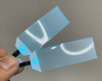 May include: Two small, rectangular, translucent blue light panels with a grid pattern. The panels are connected to a black wire with a red and black wire leading to a battery pack and a black switch.