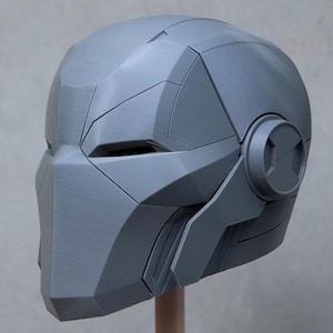 May include: A gray, futuristic-style helmet with angular panels and a circular detail on the side. The helmet is mounted on a wooden dowel, showcasing its design and structure. The overall aesthetic suggests a sci-fi or cosplay theme.