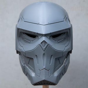 May include: A gray, futuristic-looking helmet with a detailed, angular design. The helmet features eye openings and a segmented face covering, suggesting a high-tech or sci-fi theme. It is likely a cosplay or prop item.