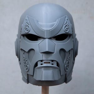 May include: A gray, 3D-printed helmet with intricate details. The helmet features a faceplate with eye openings, a nose piece, and decorative elements. The design is symmetrical and appears to be a cosplay or costume piece.