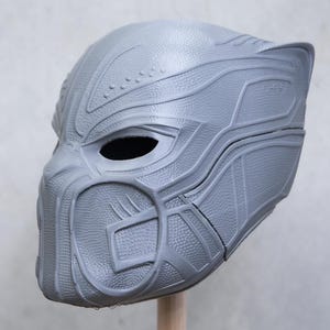 May include: A grey, 3D-printed mask with a detailed design, textured surfaces, and defined lines. The mask has eye openings and a prominent facial structure, resembling a superhero helmet. The item is mounted on a wooden dowel.