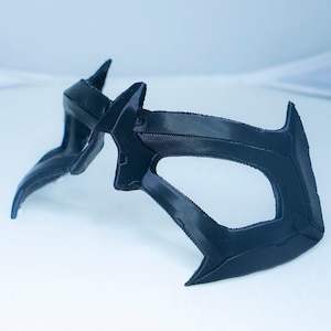 Könnte beinhalten: Eine schwarze Plastikmaske mit geometrischem Design. Die Maske hat zwei Augenlöcher und ein spitzes Kinn.