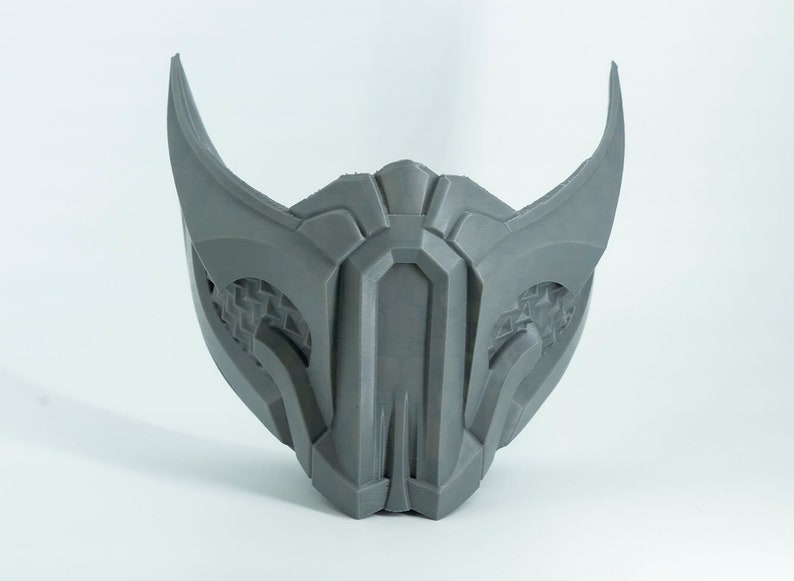 May include: A gray 3D printed mask with a geometric design and sharp points. The mask has a detailed pattern on the sides and a central ridge.