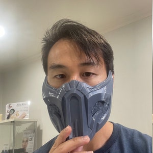 May include: A person wearing a gray 3D printed mask with a detailed design. The mask covers the person's nose and mouth. The mask is similar to the one worn by the character Sub-Zero in the Mortal Kombat video game series.