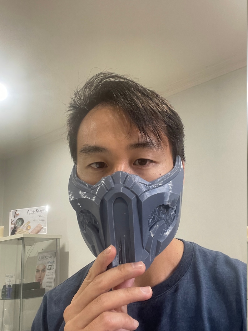 May include: A person wearing a gray 3D printed mask with a detailed design. The mask covers the person's nose and mouth. The mask is similar to the one worn by the character Sub-Zero in the Mortal Kombat video game series.