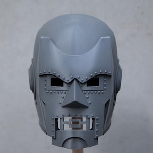 May include: A gray, full-face mask with a distinctive design, featuring rectangular eye openings, a triangular nose, and a mouth area with a grid-like pattern. The mask is adorned with numerous small, round rivets and has a rounded top.