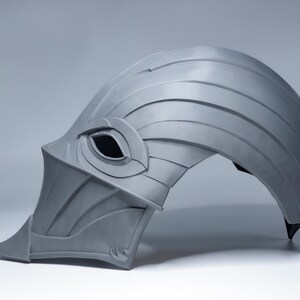 May include: A gray helmet with a pointed front and a single eye hole. The helmet is made of a textured material and has a curved shape.