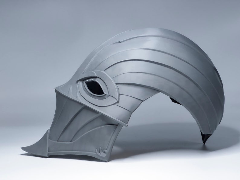 May include: A gray helmet with a pointed front and a single eye hole. The helmet is made of a textured material and has a curved shape.