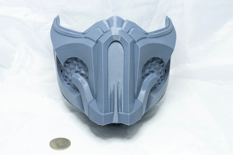 May include: A gray 3D printed mask with a detailed design. The mask has a raised, textured pattern on the front and a smooth, rounded shape.
