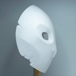 Op de afbeelding: Een witte, abstracte sculptuur die lijkt op een masker of gezicht, gemonteerd op een houten deuvel. De sculptuur heeft een gladde, afgeronde vorm met een grote, ovale opening en een getextureerde, gebroken rand. De achtergrond is effen lichtgrijs.