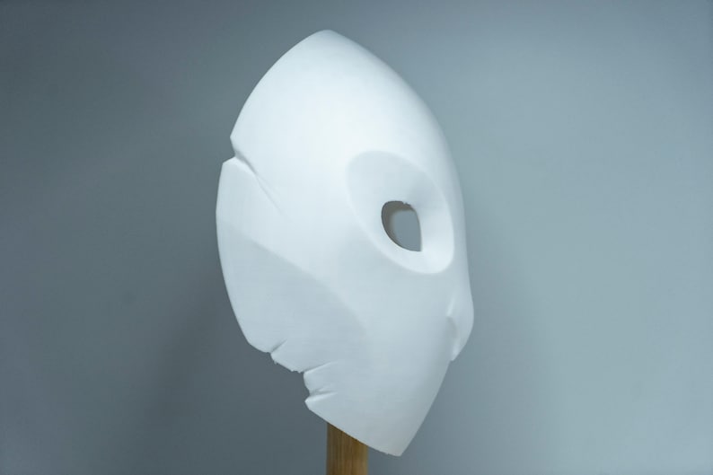 Op de afbeelding: Een witte, abstracte sculptuur die lijkt op een masker of gezicht, gemonteerd op een houten deuvel. De sculptuur heeft een gladde, afgeronde vorm met een grote, ovale opening en een getextureerde, gebroken rand. De achtergrond is effen lichtgrijs.
