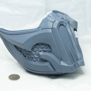 May include: A gray 3D printed mask with a detailed design. The mask has a textured surface and a sharp, angular shape.