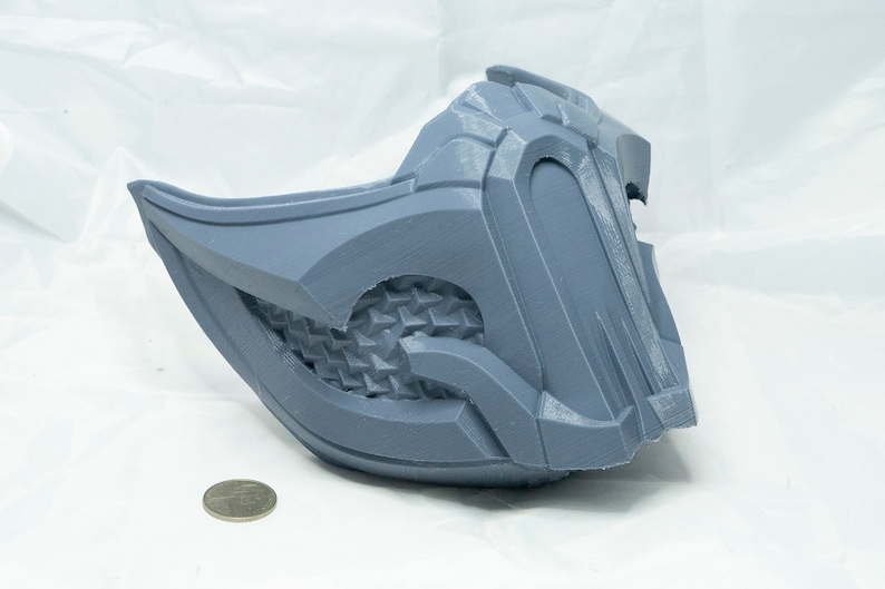 May include: A gray 3D printed mask with a detailed design. The mask has a textured surface and a sharp, angular shape.
