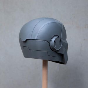 May include: A gray, futuristic-looking helmet with a rounded top and angular features. The helmet has a circular detail on the side and is mounted on a wooden dowel. The design suggests a sci-fi or cosplay theme.