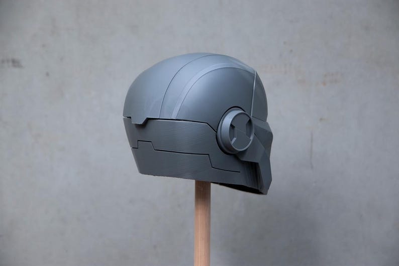 May include: A gray, futuristic-looking helmet with a rounded top and angular features. The helmet has a circular detail on the side and is mounted on a wooden dowel. The design suggests a sci-fi or cosplay theme.